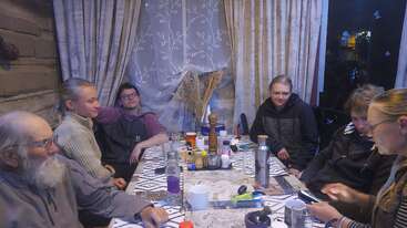 Seis personas se sientan alrededor de una mesa cubierta de tazas, botellas y utensilios, compartiendo un momento acogedor en el interior. Parecen relajados, hablando juntos en una habitación cálidamente iluminada.