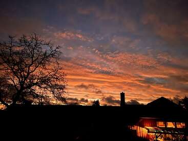 Un coucher de soleil spectaculaire emplit le ciel de nuages orange et roses éclatants. La silhouette d'un arbre et une maison chaleureuse éclairée par des lumières douces créent une scène paisible en soirée.