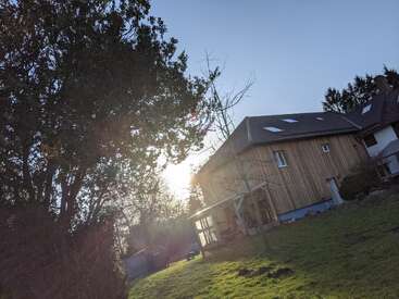 L'image montre une maison en bois avec un toit en pente et une cour avec de l'herbe verte, des arbres et la lumière du soleil qui brille à travers les branches, créant une atmosphère paisible.