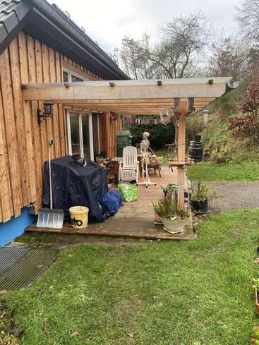 Auf der Terrasse eines Holzhauses stehen Stühle, Topfpflanzen, zugedeckte Sachen, ein Plastikskelett am Tisch, bunte Fahnen und ein grasbewachsener Garten an einem bewölkten Tag.