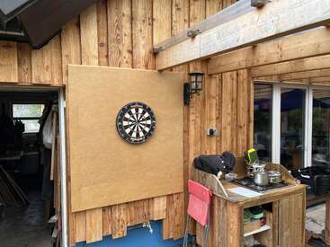 A dartboard is mounted on a wooden board outside a rustic wooden house. Next to it, there’s an outdoor stove with a pot, chair, and kitchen items.