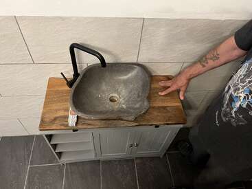 A unique stone sink sits atop a wooden vanity with open shelves, black faucet, and tile wall. A tattooed person’s hand rests on the counter.