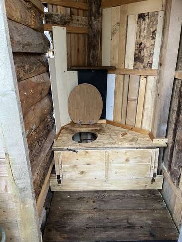 This image shows a rustic, wooden outdoor toilet or outhouse with a simple wooden seat, surrounded by wooden planks and logs, creating a primitive, natural look.
