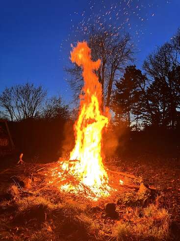 A large, bright bonfire burns intensely, sending flames and sparks upward. The fiery blaze contrasts against a deep blue evening sky with silhouetted trees surrounding it.