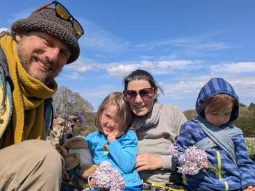 A happy family of four sits outdoors on a sunny day. They’re surrounded by wildflowers, with two children holding bunches. Everyone is smiling and enjoying nature.