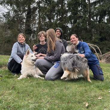 Five people and two dogs sit together on grass, smiling and enjoying each other's company in an outdoor setting with tall trees in the background, appearing happy.