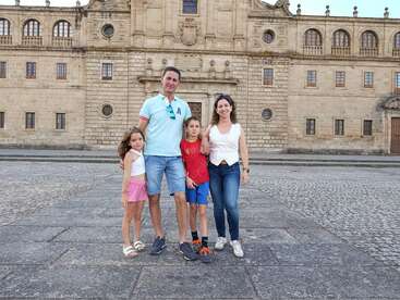 Uma família de quatro pessoas posa junta em frente a um grande edifício histórico de pedra, sorrindo, aproveitando um dia claro, vestida casualmente com roupas de verão.