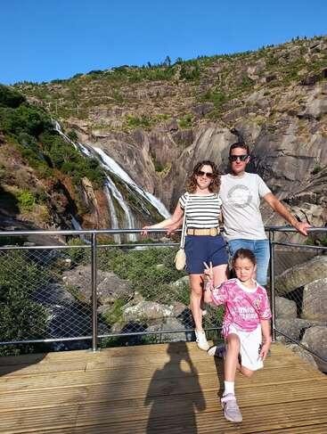 Uma família de três pessoas posa em uma plataforma de madeira com uma cachoeira exuberante e colinas rochosas ao fundo. A criança na frente faz um sinal de paz.