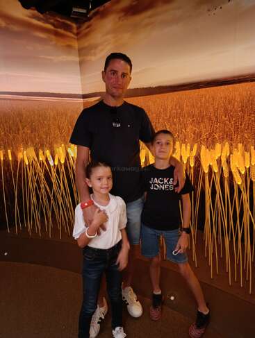 Um homem e duas crianças posam para uma foto em frente a um pano de fundo que mostra um campo de trigo dourado sob um céu nublado e dramático ao pôr do sol.