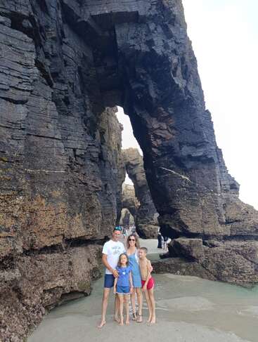 Uma família de quatro pessoas posa alegremente em uma praia de areia, cercada por formações rochosas imponentes e dramáticas, com arcos naturais e superfícies texturizadas e ásperas. Belo cenário.