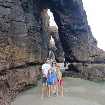 A family of four poses happily on a sandy beach, surrounded by dramatic, towering rock formations with natural arches and textured, rugged surfaces. Beautiful scenery.