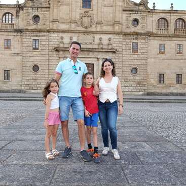 A family of four poses together in front of a large historic stone building, smiling, enjoying a bright day, dressed casually in summer clothes.