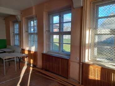 This image shows a classroom with large windows protected by wire mesh, wooden radiators, scattered desks, and sunlight streaming through, illuminating the worn, empty room.