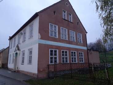 This image shows an old, two-story house with many windows, faded beige and white walls, surrounded by a fence and located on a quiet street.