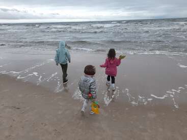 Three children in jackets walk toward a cold, windy beach. Waves crash gently, and the sky is overcast. They carry colorful beach toys, exploring together.