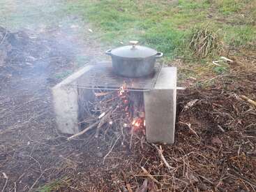 A black pot sits on a grill supported by two cinder blocks, with a fire made from sticks burning underneath, outdoors on grass and dirt.