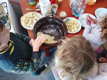 Drei Kinder sitzen um einen Tisch herum und formen aus Teig kleine Kugeln. Auf dem Tisch liegen Teller mit Teigkugeln, Mehl, Tassen und Küchenutensilien verstreut.