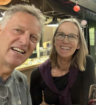 Um homem e uma mulher sorridentes sentados juntos em um ambiente interno, possivelmente em um restaurante ou café, com iluminação quente, bolos atrás deles e uma taça de vinho tinto.