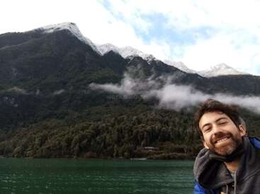 A man smiles at the camera in the foreground, while behind him lies a scenic view of green forested mountains, snowy peaks, and a calm lake.