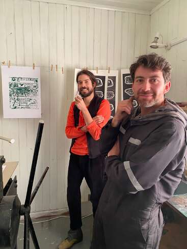 Two men stand in an art studio, smiling and posing thoughtfully. Artwork hangs behind them, and the space has white wood-paneled walls and art supplies.