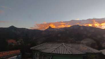 Um pôr do sol deslumbrante ilumina as nuvens atrás de picos montanhosos escarpados, com um cume pontiagudo bem no centro. Em primeiro plano, telhados de telhas e prédios conferem textura e profundidade.