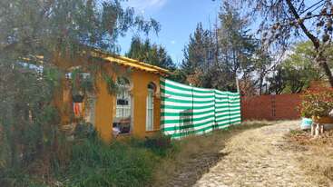 Uma casinha amarela com janelas em arco fica ao lado de um gramado cheio de mato. Uma cerca de lona listrada de verde e branco acompanha um caminho de paralelepípedos, cercado por árvores.