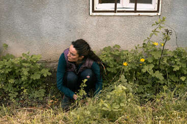 Une femme aux longs cheveux bruns est accroupie près d'un mur, entourée de plantes vertes et de fleurs jaunes ; on dirait qu'elle jardine ou qu'elle examine attentivement les mauvaises herbes.