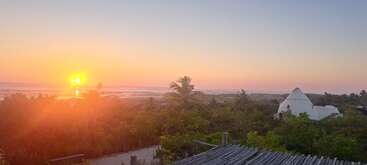 Un magnifique coucher de soleil sur l'océan, des palmiers, une végétation luxuriante et un bâtiment blanc en forme de pyramide créent une atmosphère paisible, tropicale et sereine.