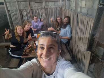 Un groupe de six amis est assis autour d'une table rustique en bois à l'intérieur d'une pièce aux murs de bambou, souriant et posant pour un selfie joyeux, profitant du temps qui leur est imparti.