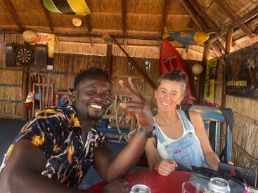 Un homme et une femme souriants sont assis ensemble dans un bar rustique au toit de chaume, décoré d'affiches, d'un jeu de fléchettes et d'objets colorés. Ils prennent un verre et discutent en toute décontraction.