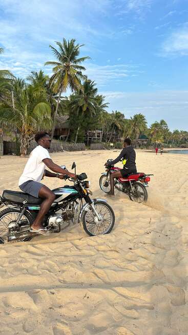 Deux hommes roulent à moto sur une plage de sable bordée de palmiers, sous un ciel d'un bleu éclatant. L'ambiance tropicale, la détente et l'aventure se mêlent ici de manière harmonieuse.