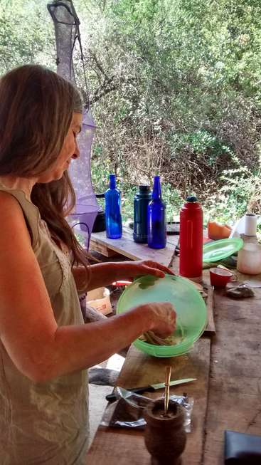 A imagem mostra uma mulher preparando comida em um ambiente rústico ao ar livre, com uma mesa de madeira e vários itens, incluindo garrafas e uma garrafa térmica vermelha, ao fundo.