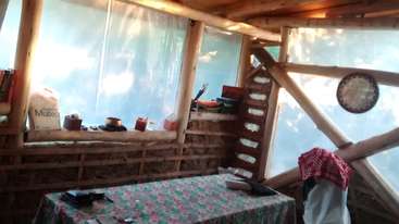 A imagem mostra uma sala com uma mesa e cadeiras, com uma grande janela no lado esquerdo. As paredes são pintadas de branco e o piso é de madeira. A atmosfera geral é clara e arejada.