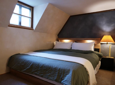 Cette chambre confortable présente un plafond incliné, un grand lit en bois avec des draps verts, des oreillers blancs moelleux, une lampe de chevet et une fenêtre qui laisse entrer la lumière.