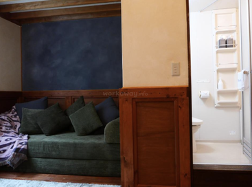La chambre confortable comprend un canapé vert avec plusieurs coussins, des panneaux en bois, une couverture et une salle de bains moderne adjacente avec des toilettes et des étagères encastrées.