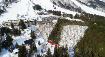 Vue aérienne d'un paysage enneigé comprenant des bâtiments, des forêts de pins denses, un parking, des pistes de ski et des arbres couverts de neige dans une station de montagne.