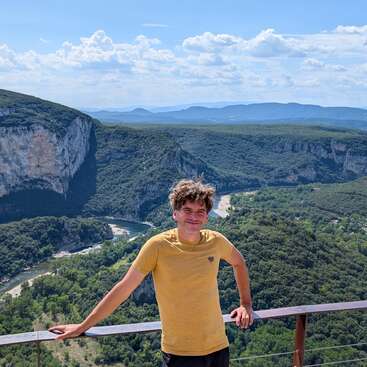 Un homme souriant, vêtu d'une chemise jaune, se tient près d'une balustrade, surplombant une superbe vallée verte avec une rivière sinueuse, des falaises et de lointaines montagnes bleues sous un ciel lumineux.