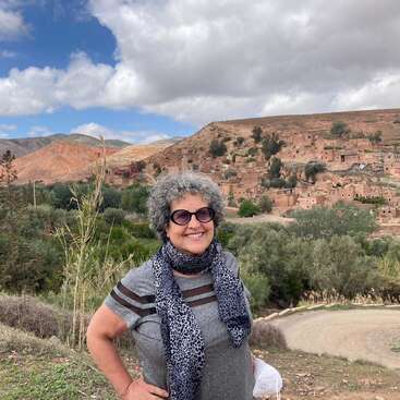 Una mujer alegre, con el pelo gris y rizado y gafas de sol, posa al aire libre, vestida con un pañuelo y una camiseta. Detrás de ella se ven unas casas rústicas en la ladera y una exuberante vegetación.
