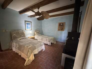 This cozy bedroom features two twin beds with patchwork quilts, exposed wood beams, a ceiling fan, wood stove, blue walls, brick floor, and soft lighting.