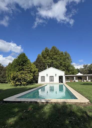 A charming white house stands at the end of a rectangular swimming pool, surrounded by lush green grass, trees, and a bright, partly cloudy sky.
