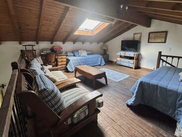 This cozy attic room features wooden ceilings, two blue beds, a sofa with striped cushions, a coffee table, TV, skylight, books, and charming rustic décor.