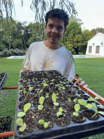 A smiling man holds a tray of young seedlings outdoors. He stands on a grassy lawn, with trees, a pool, and a white house in the background.