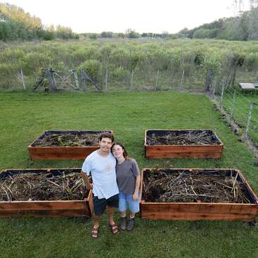 A couple stands in front of four large wooden raised garden beds in a grassy backyard, surrounded by lush green fields and a rustic wire fence.