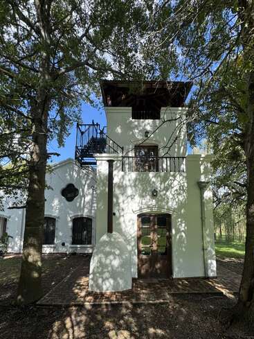 Ein charmantes weißes zweistöckiges Haus mit Holztüren und einer Außentreppe, umgeben von Bäumen, deren Schatten auf die Fassade fallen, in einer ruhigen, natürlichen Umgebung.