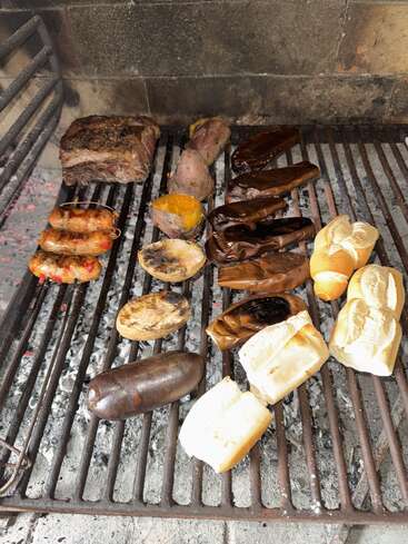 Ein Grill mit verschiedenen Fleischsorten, Würstchen, Blutwurst, Brötchen, Süßkartoffeln, geschnittenem Gemüse und gebratenen Auberginen über glühender Kohle in rustikaler Atmosphäre.