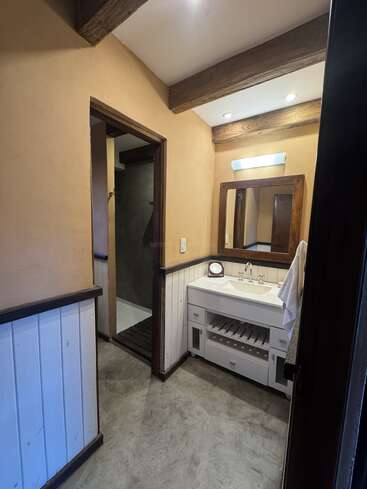Cette photo montre une salle de bains au style rustique, avec des poutres apparentes, un meuble-lavabo, un miroir, des murs dans des tons beiges chauds et un espace douche adjacent partiellement visible.