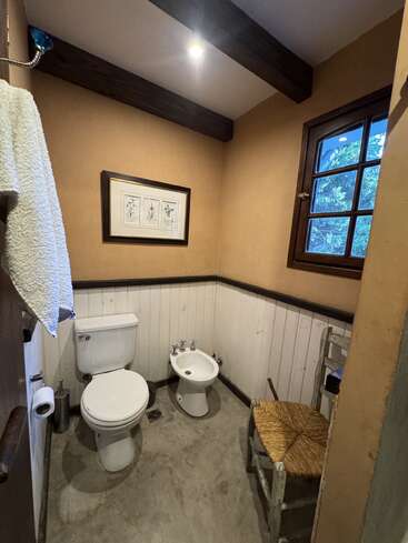 Cette salle de bains chaleureuse comprend des toilettes, un bidet, une chaise rustique en bois, une serviette, une reproduction botanique encadrée, des poutres apparentes, un éclairage tamisé, une fenêtre et des murs peints en beige avec des demi-lambris.