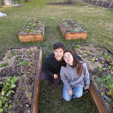 A couple kneels closely together in a garden, surrounded by four raised wooden beds with various plants, enjoying a peaceful, sunny day outdoors.