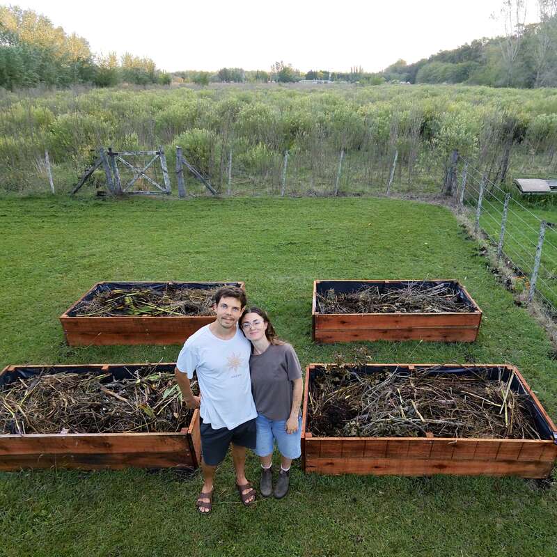 A couple stands in front of four large wooden raised garden beds in a grassy backyard, surrounded by lush green fields and a rustic wire fence.