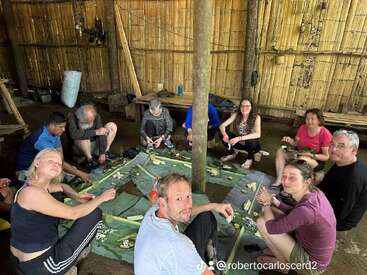 Um grupo de pessoas está sentado em círculo no chão, dentro de uma cabana rústica de bambu, preparando comida juntos, usando folhas de bananeira e vários ingredientes, curtindo essa atividade em comunidade.